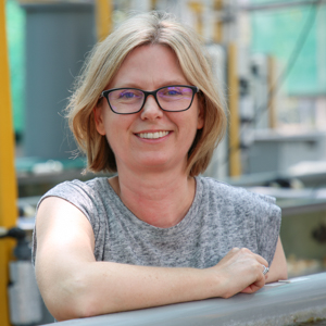 Woman with Blond hair and glasses wearing grey shirt, standing in front of equipment