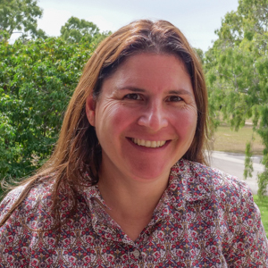 Woman with brown hair wearing patterned shirt standing in front of trees.