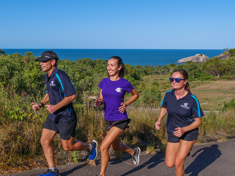 three people are running along a trail with bush and ocean in the background