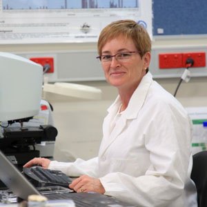 Woman standing at a computer, wearing reading glasses and a white laboriatory coat.