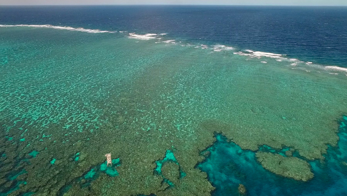 A photo from the air of a large reef platform at high tide, with blue water and the horizon in the distance. A white metal tower stands on the reef top.