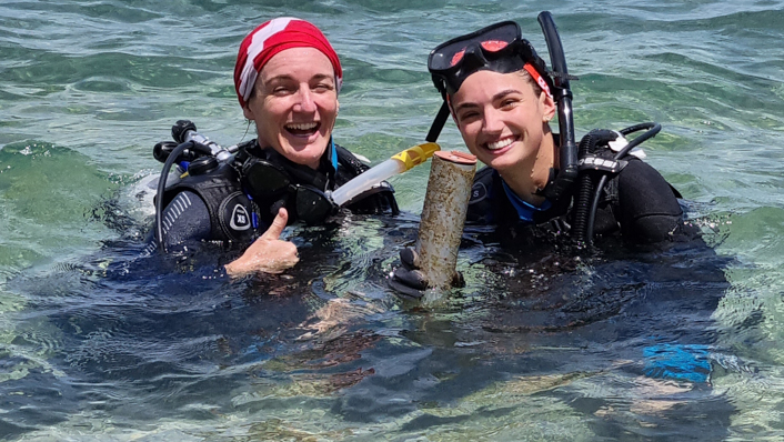 Two women in scuba diving gear are at the surface holding data logging equipment