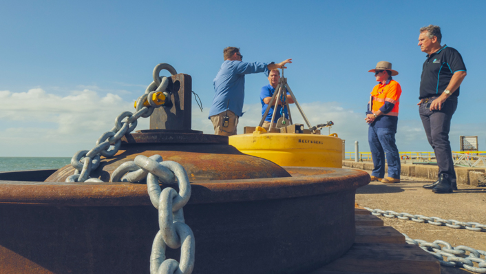 An anchor and buoy sit on a wharf whilst people in the background talk