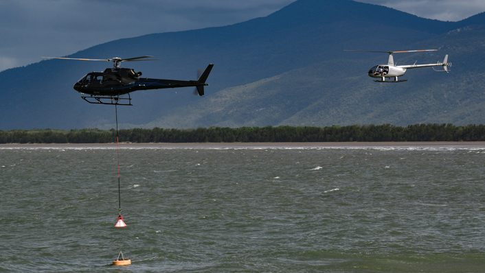 A helicopter has a chain daggling from underneath as it retrieves a buoy at sea. Another helicopter hovers in the background