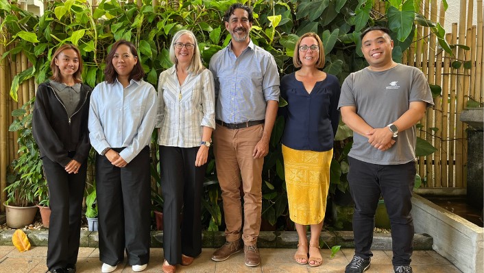 Six people smile at the camera while standing in a courtyard area