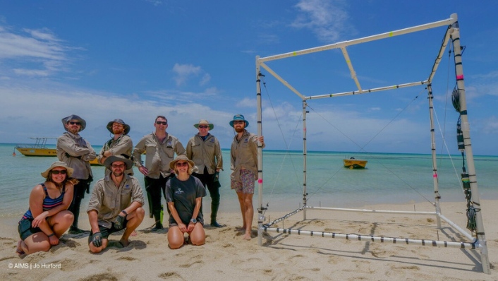 a group of people stand in a group on a beach, smiling at the camera. there is a large white frame structure to the side of them