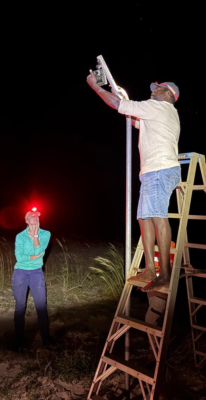 A man on a ladder is adjusting a camera set at a high level while a woman watches him from the ground