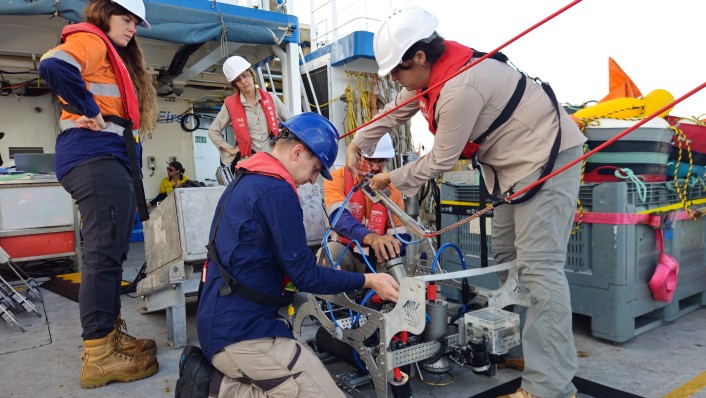 A group of people in work wear are shown working on metal framed equipment on a large boat deck