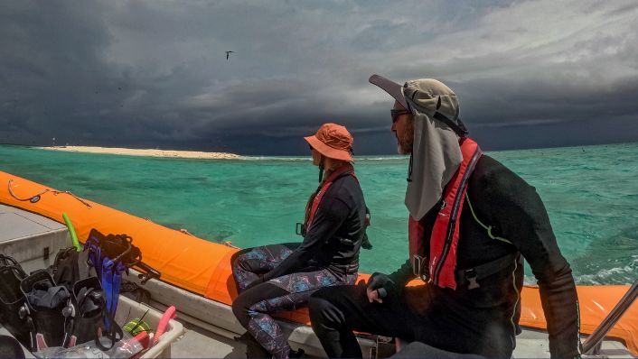 Photo of two reef survey team members seated in a small boat beside a coral reef cay, watching a dark storm front approach across turquoise water. Dive gear is visible in the boat, illustrating fieldwork undertaken in challenging weather conditions on the Great Barrier Reef.