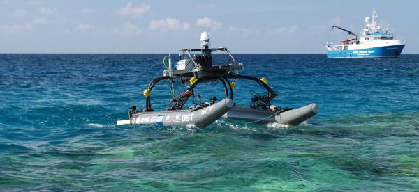 grey and black marine robot on the surface of the ocean with blue and white AIMS research vessel in the distane
