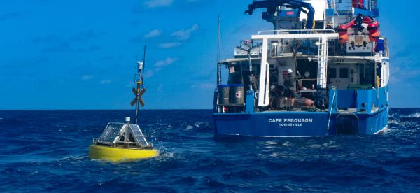 a yelloe ocean buoy sits in the water with the stern of the Cape Ferguson research vessel behind it.