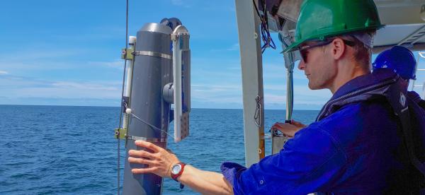 A scientist with hard hat with a large grey bottle over the side of a large vessel
