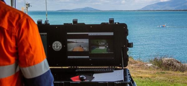 looking over a person shoulder wearting high vis shirt, and seeing a computer screen in front of them. IN the distance there is calm ocean water and a small floating robot at the surface.