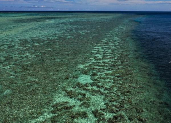 a low aerial photo of a reef flat at high tide dropping off to deeper water on the right of the photo. A small orange boat with a person in it is over the deeper blue water.
