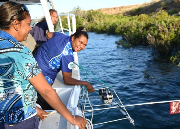 Two Indigenous Rangers lean over the side of a small boat in mangroves whilst deploying a baited remote underwater video station