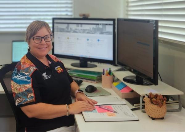 A person, smiling, sitting at a desk at home with computer screens and papers on the desk