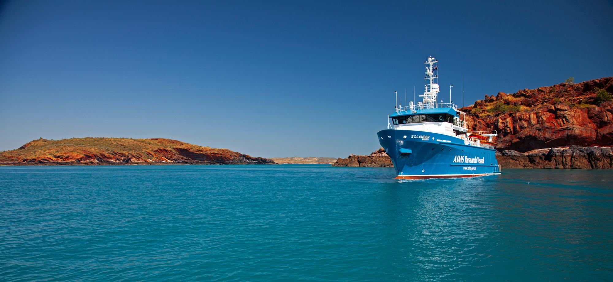 blue and white research vessel on water with orange dirt cliff behind