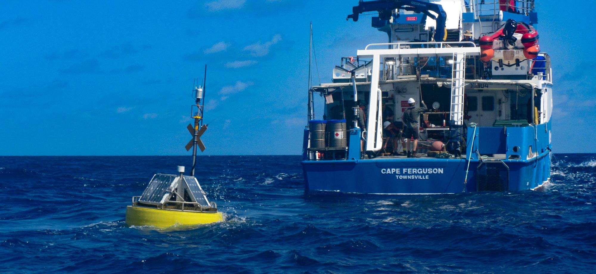 a yelloe ocean buoy sits in the water with the stern of the Cape Ferguson research vessel behind it.