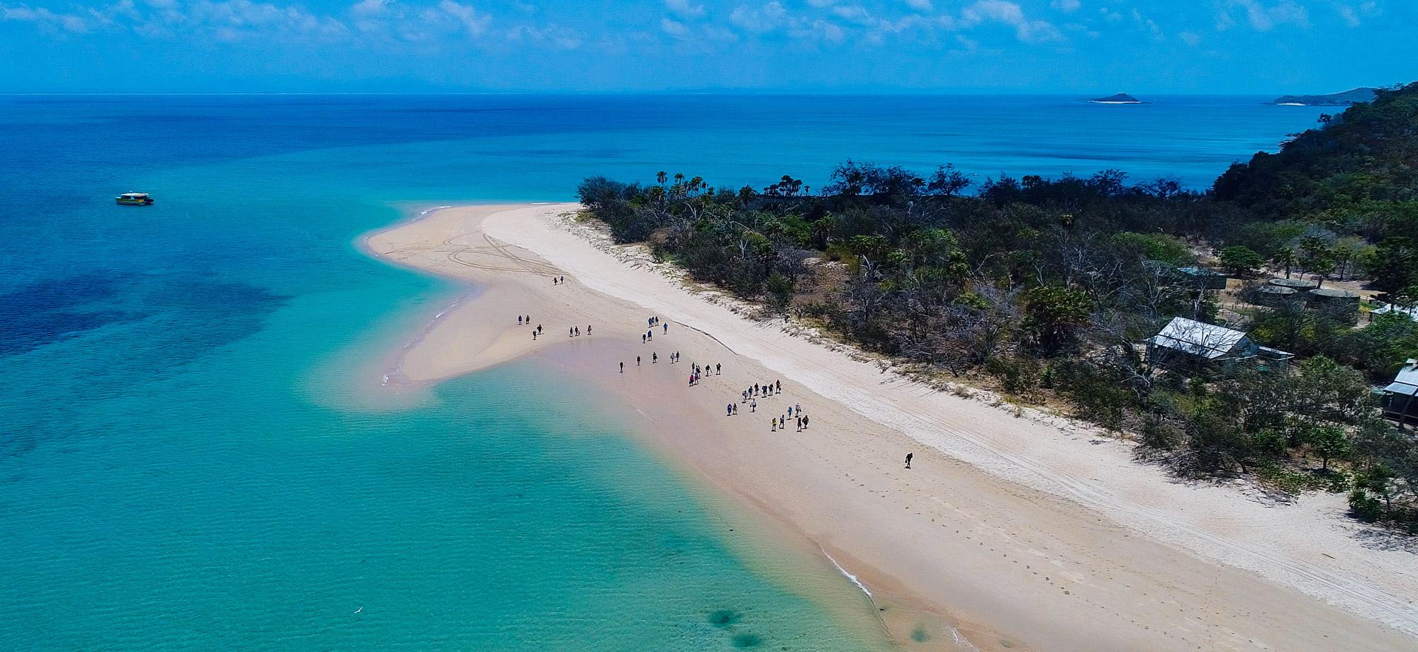 aerial view over white sand beach with people walking on it, next to a turquoise waters of the ocean