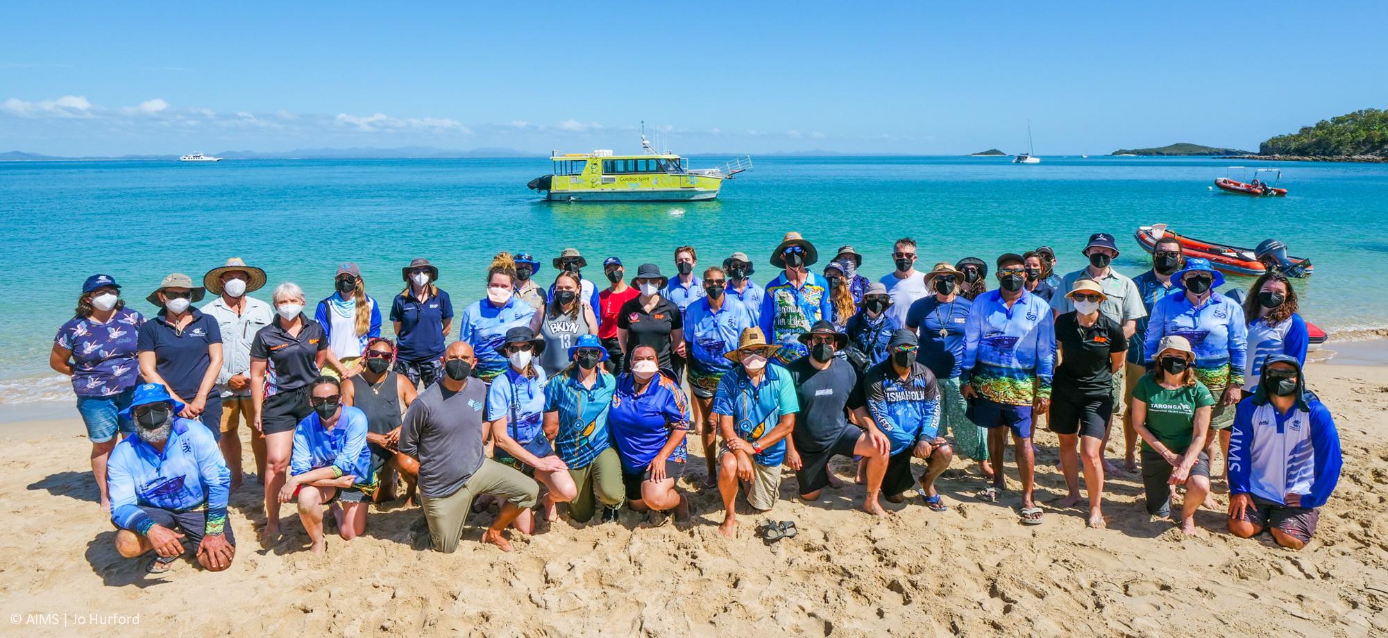many people in masks grouped on a beach looking at the camera with a calm tropical sea in the background, and a boat