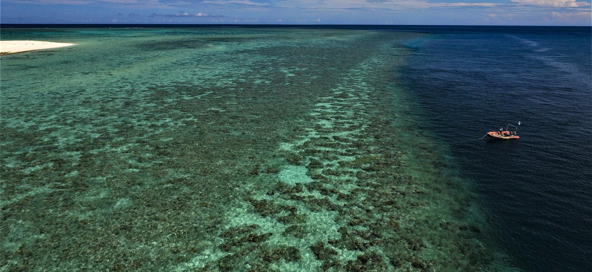 a low aerial photo of a reef flat at high tide dropping off to deeper water on the right of the photo. A small orange boat with a person in it is over the deeper blue water.