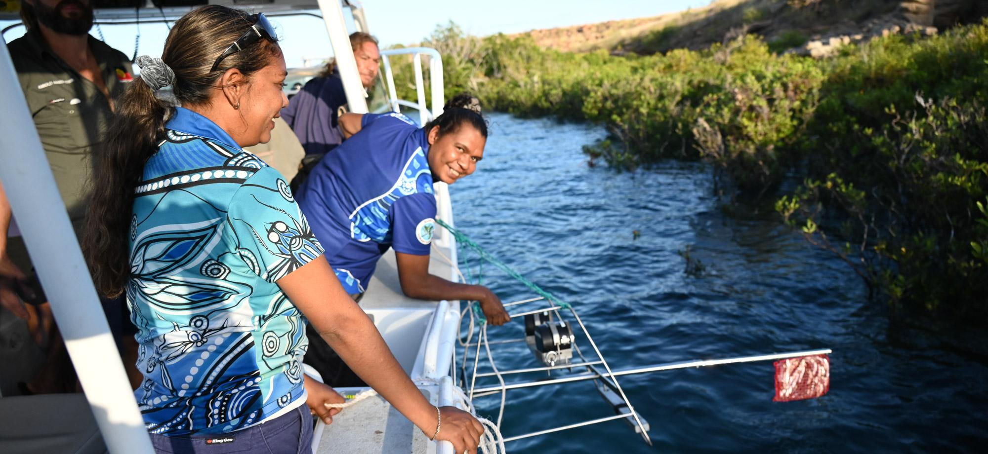 Two Indigenous Rangers lean over the side of a small boat in mangroves whilst deploying a baited remote underwater video station