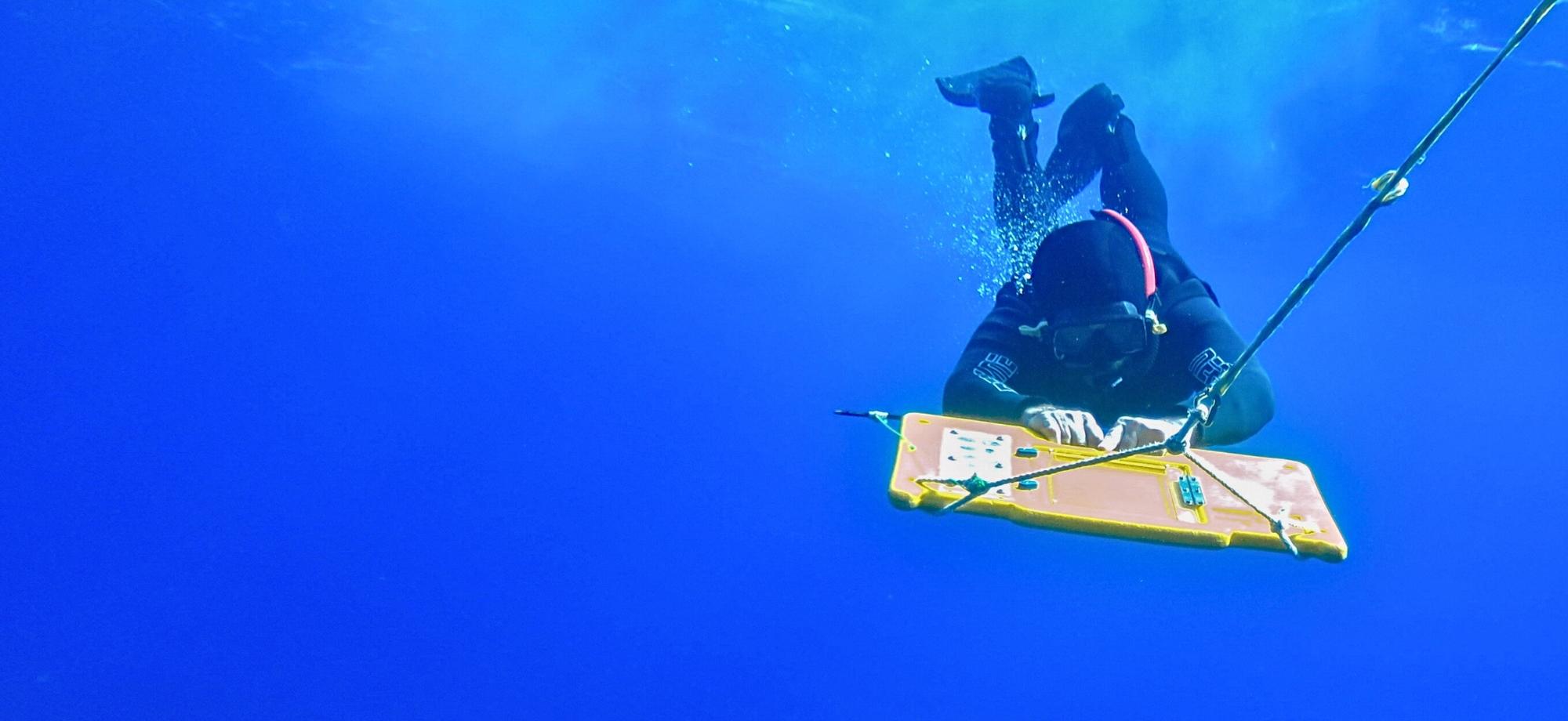 Underwater scene showing a diver in a black wetsuit and mask holding a yellow rectangular device connected to a rope, with bubbles rising in deep blue water