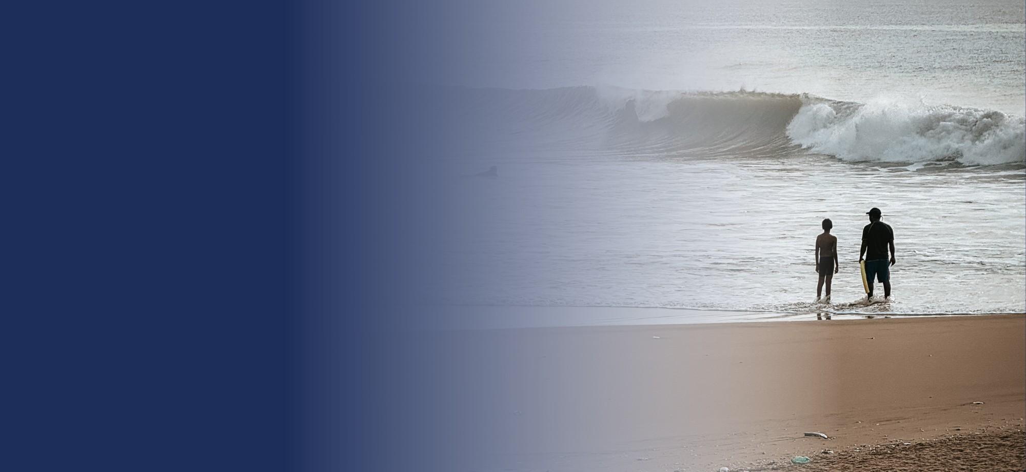 silhouettes of 2 people standing in the shallows of a beach with a small wave in the background. it is early morning or late afternoon.