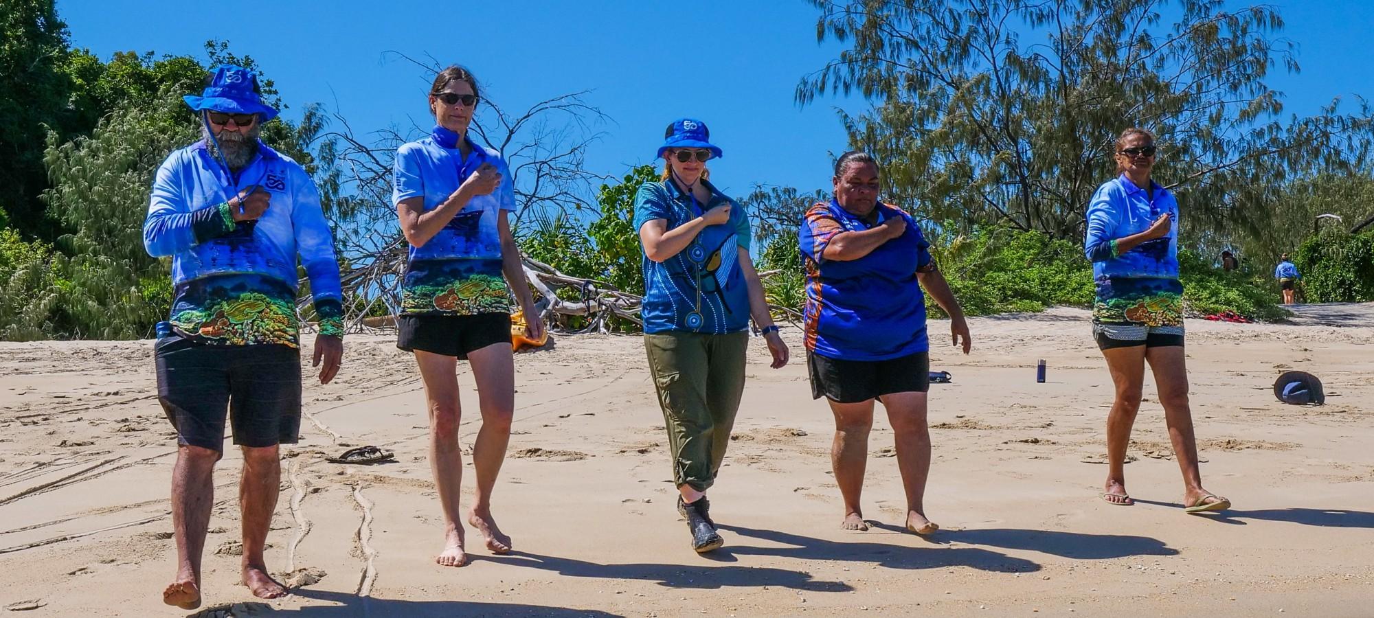 Five people in matching blue project shirts walk barefoot along the shoreline, each with one hand placed on their chest, with coastal vegetation behind them and shallow waves at their feet.