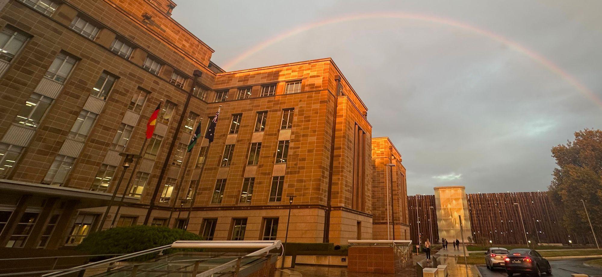 Photo of a sandstone government building in Canberra bathed in golden sunset light after rain, with national flags at the entrance. A double rainbow arches across a grey sky above the building, while wet pathways, a few vehicles and pedestrians are visible in the foreground.