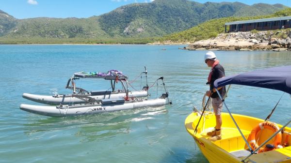 A person in a hard hat stands on a small yellow boat looking at a twin hull, grey vessel that has no people on board