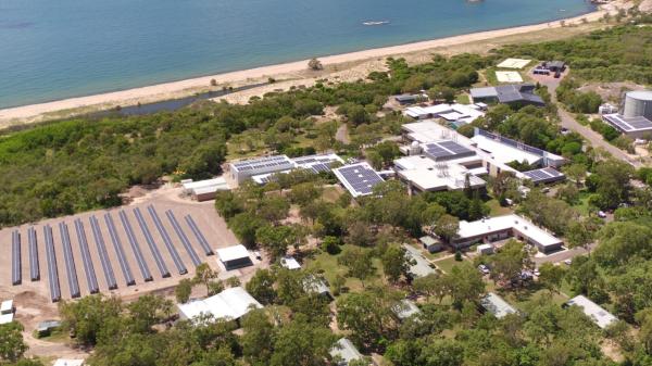 This photo is taken from the air. Buildings with solar panels are surround be green grass and bush. The ocean and beach can be seen at the top of the photo. 