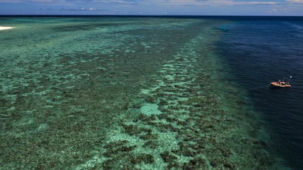 a low aerial photo of a reef flat at high tide dropping off to deeper water on the right of the photo. A small orange boat with a person in it is over the deeper blue water.