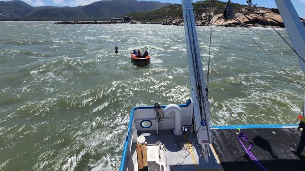 a view over the stern of a vessel at small orange inflatable in the water, and a green, hilly coastline in the distance