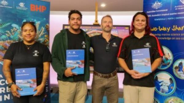 4 people stand smiling at the camera in front of promotional banners. 3 of the people are holding certificates