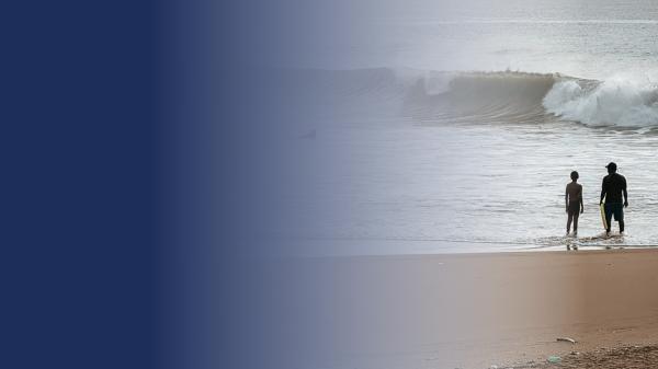 silhouettes of 2 people standing in the shallows of a beach with a small wave in the background. it is early morning or late afternoon.
