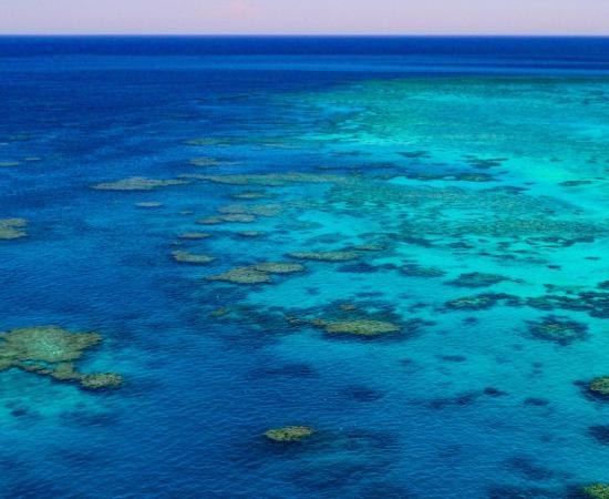 Aerial photo of the Great Barrier Reef showing coral reef formations and shallow turquoise lagoons surrounded by deep blue ocean. A small boat is visible beside one of the reef platforms, highlighting the scale of the reef landscape.