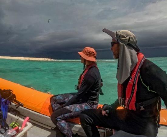 Photo of two reef survey team members seated in a small boat beside a coral reef cay, watching a dark storm front approach across turquoise water. Dive gear is visible in the boat, illustrating fieldwork undertaken in challenging weather conditions on the Great Barrier Reef.