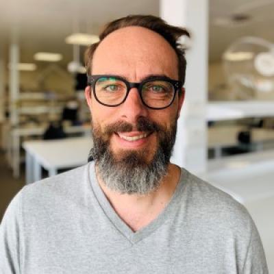 Bokeh effect image of a bearded man with dark brown hair, wearing glasses and a grey shirt with out-of-focus work-space background.