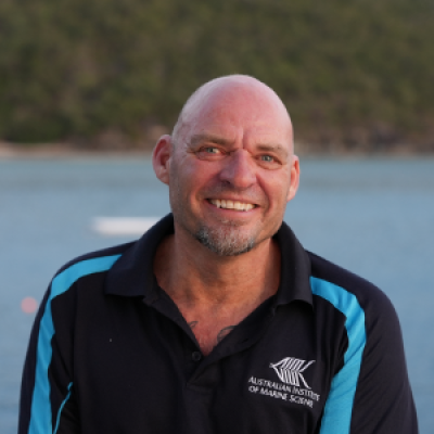 Man wearing blue and black AIMS shirt, with an ocean and green bushy mountain background.