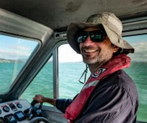 person with hat simling at camera while driving a small boat with a half cabin